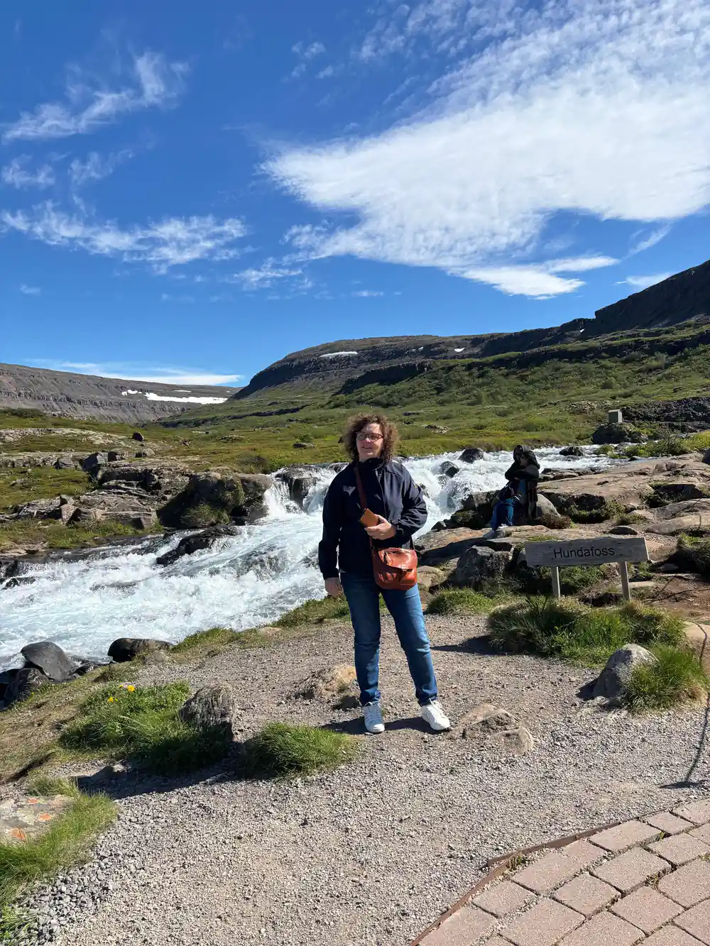 A visitor standing on the path beside the river under a blue sky