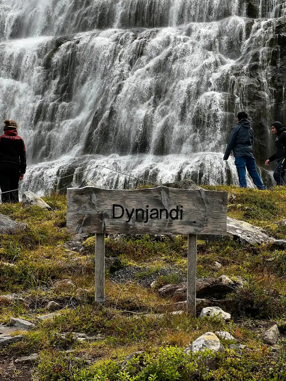 The wooden Dynjandi sign in front of a wide lower fall with people beside it